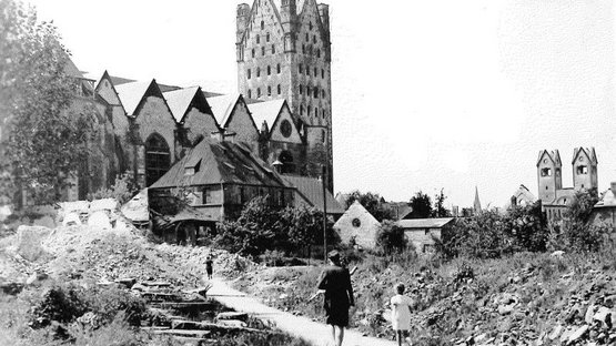 Das Foto von 1949 zeigt im Vordergrund eine Mutter mit Kind, die durch das durch den Zweiten Weltkrieg massiv zerstörte Paderborn auf den Dom zuläuft, der ebenfalls zum Teil zerstört wurde. 